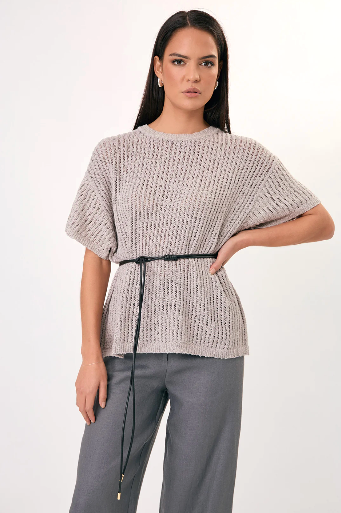 Woman wearing a textured beige top with a black belt and gray pants on a white background
