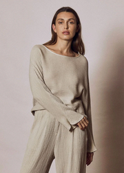 A woman wearing a beige long-sleeve top with a V-neckline and a pair of matching pants, standing against a pink background.