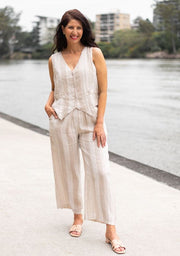 A person standing by a body of water, wearing Valentina Striped Linen Pants with a beige and white stripe pattern, paired with a white sleeveless top.