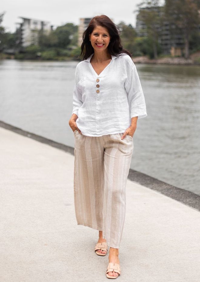 A person standing by a body of water, wearing Valentina Striped Linen Pants with a beige and white stripe pattern, paired with a white sleeveless top.