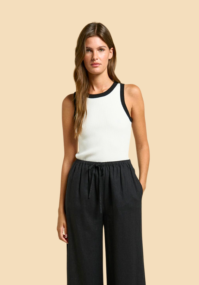 Woman wearing a white tank top with black trim and black pants on a beige background