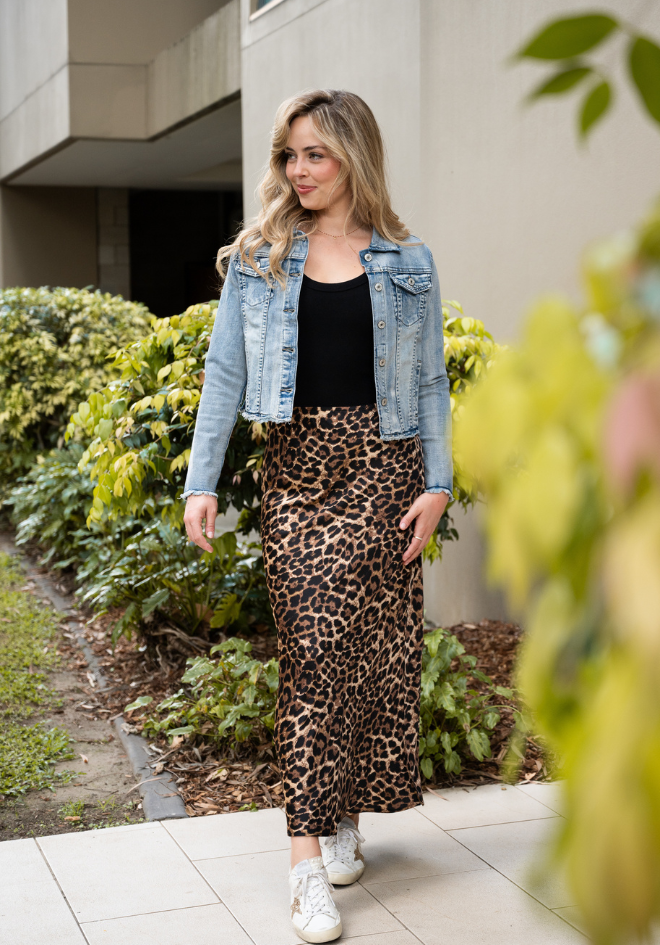 Woman wearing a denim jacket, black top, and leopard print pants standing outdoors.