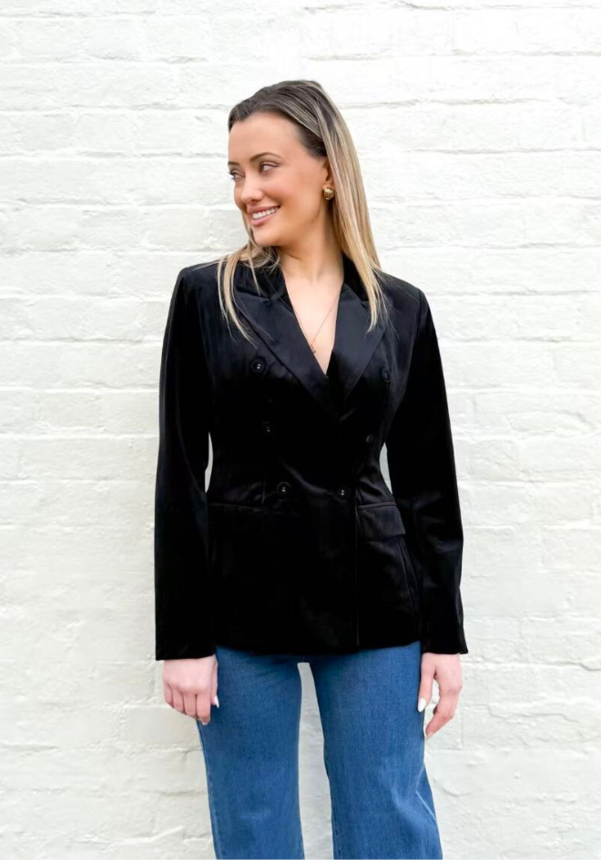 Woman wearing a black blazer and blue jeans against a white brick wall.