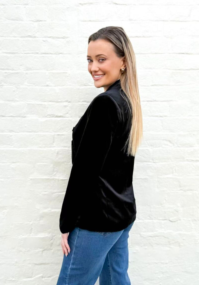Woman wearing a black velvet jacket and blue jeans against a white brick wall.