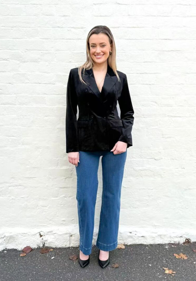 Woman wearing a black velvet blazer and blue jeans standing against a white brick wall.