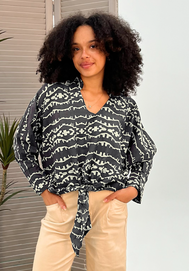 Woman wearing a black and white patterned blouse with beige pants against a neutral background