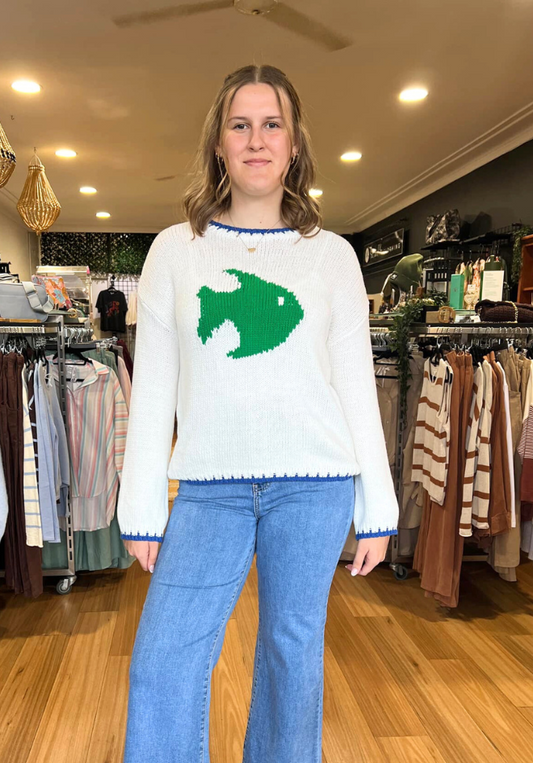 Person wearing a white sweater with a green fish design in a clothing store.