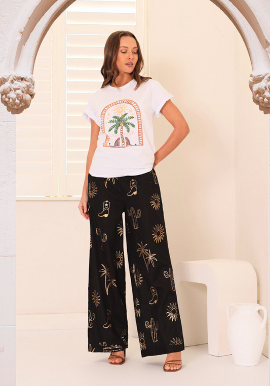Woman wearing a white t-shirt with a palm tree design and black patterned pants in a bright room.