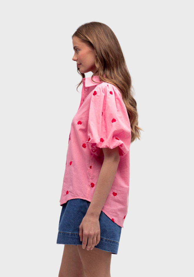 Woman wearing a pink blouse with red heart patterns and denim shorts on a white background