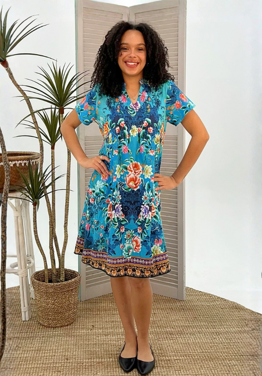 Woman wearing a colorful floral dress standing in a room with plants and a white wall.