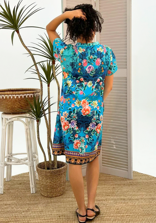 Woman wearing a blue floral dress standing in a room with a plant and stool.