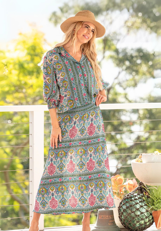 Woman wearing a patterned dress and hat on a balcony with greenery in the background
