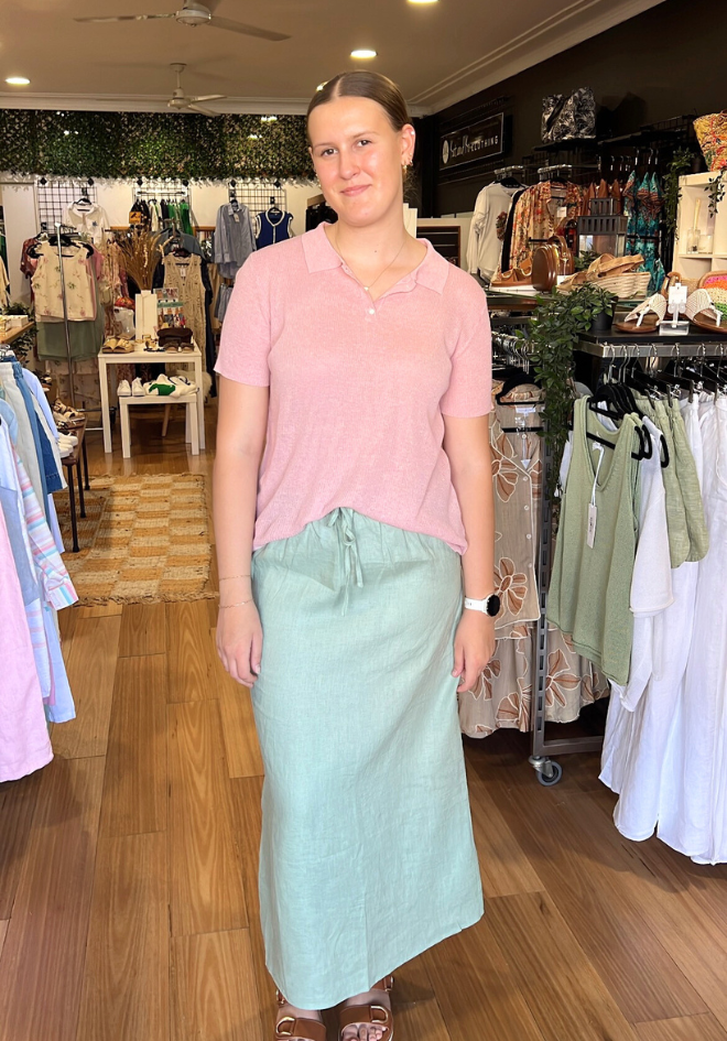 Woman standing in a clothing store wearing a pink shirt and light blue skirt.
