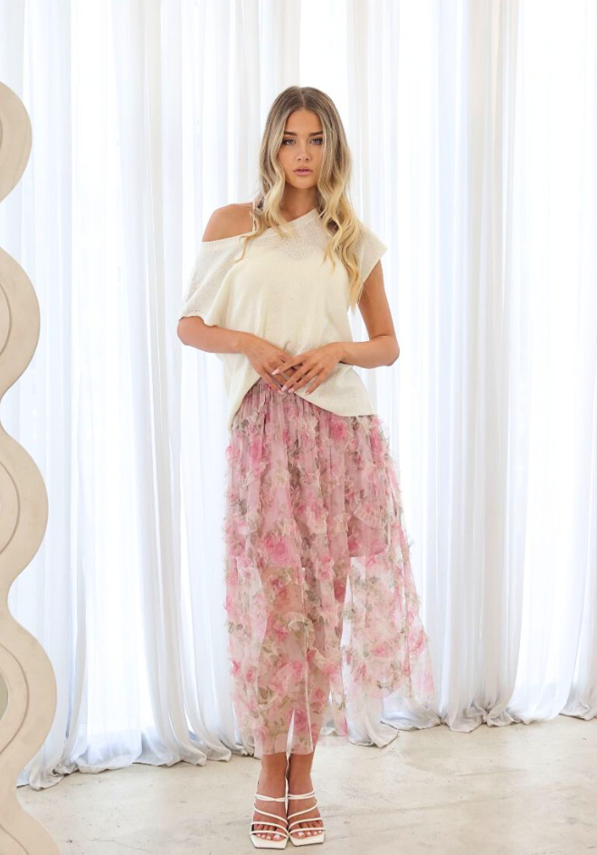 Woman wearing a white off-shoulder top and pink floral skirt against a white curtain background