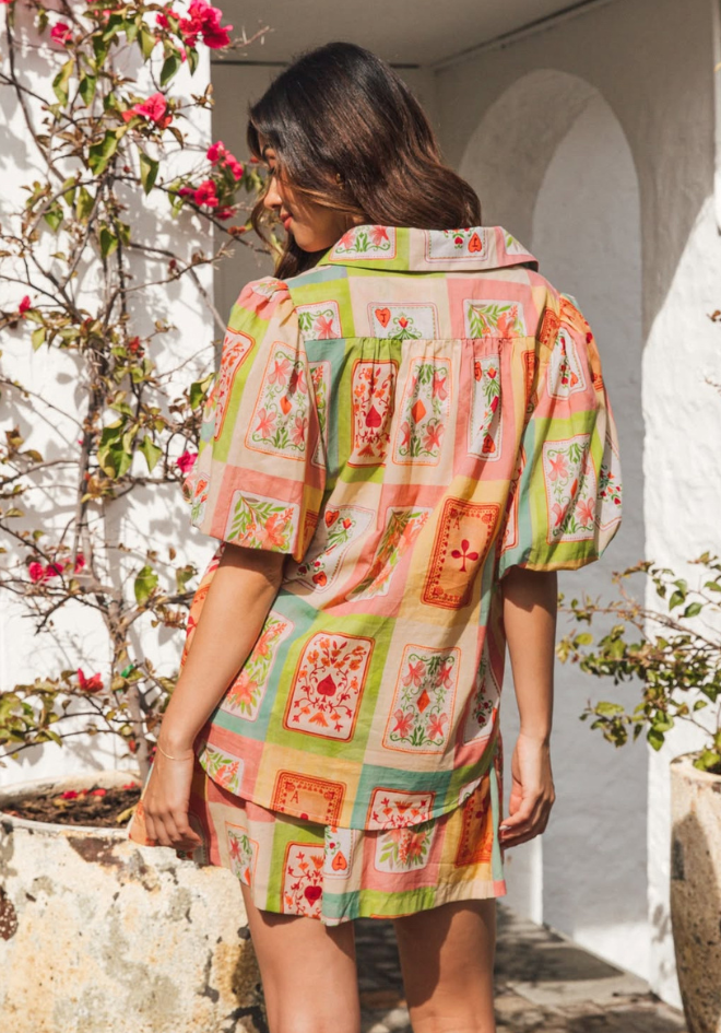 Woman wearing a colorful patterned dress standing outdoors with flowers and a white archway in the background.