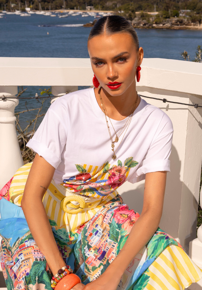 Woman sitting outdoors by a waterfront wearing a colorful skirt and white top.