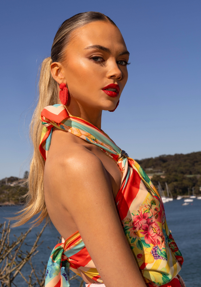 Woman in a colorful floral dress standing by a body of water with a clear blue sky.