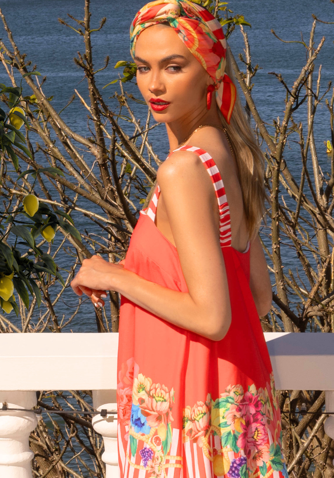 Woman wearing a red floral dress with a headscarf, standing by a body of water.