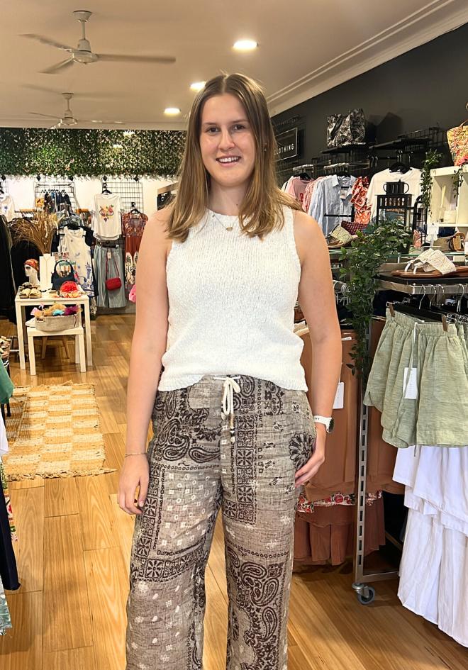 Woman standing in a clothing store wearing a white sleeveless top and patterned pants.