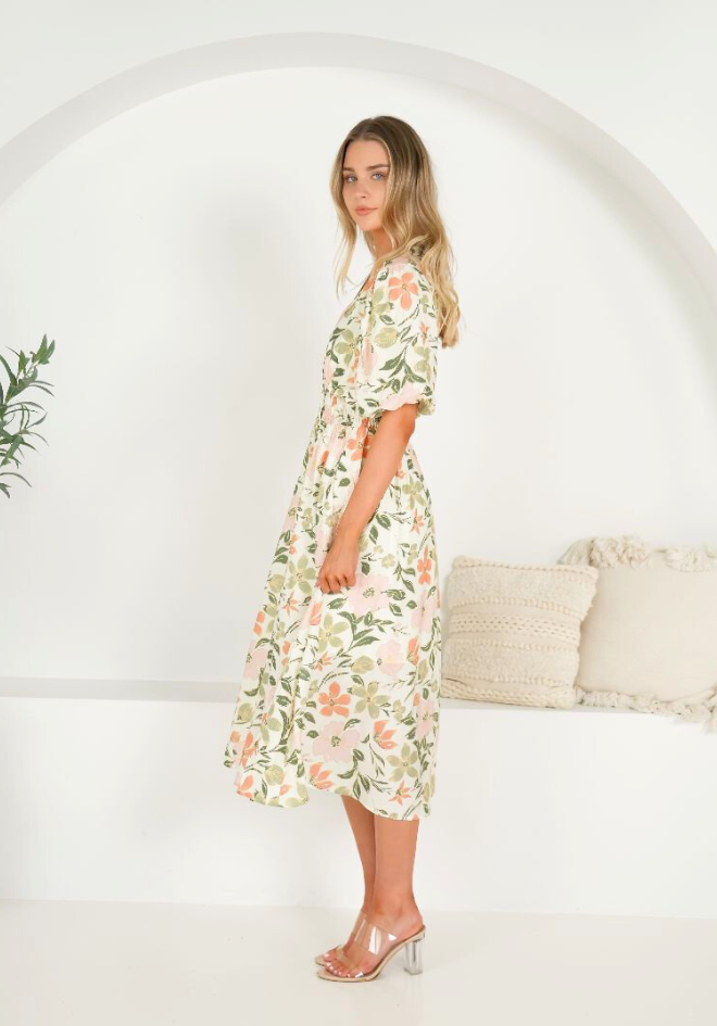 Woman wearing a floral dress standing against a white wall with a plant and cushion in the background.