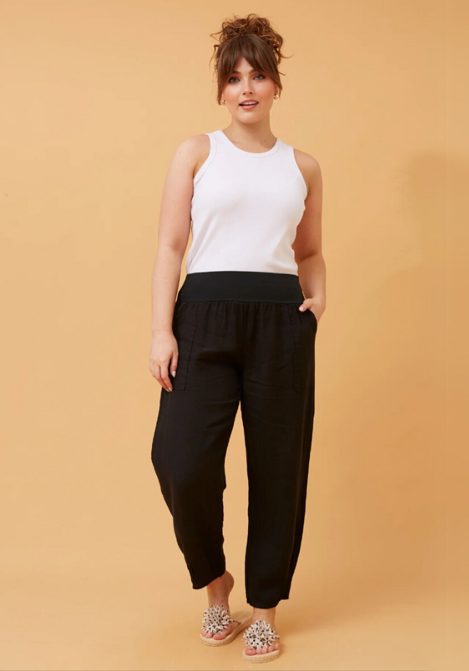 A model wearing black linen pants with a wide elastic waistband and a white top, standing against a beige background.