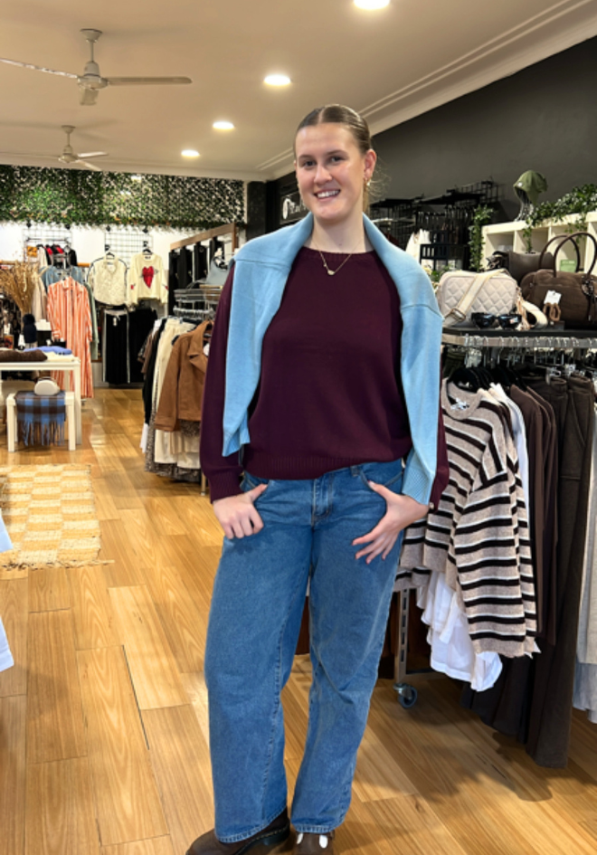Little Lies Victoria Jumper Burgundy — in-store full-length view of deep burgundy relaxed knit jumper with wide boat neckline, layered under a sky blue vest and styled with straight-leg denim jeans, available in Australia at Kat and Ko Clothing